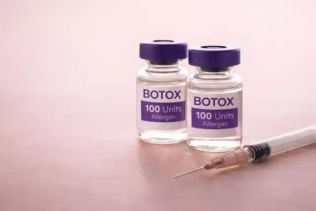 Two clear vials labeled “BOTOX 100 Units Allergan” with purple caps sit on a pink surface. In front of the vials lies a syringe with a needle pointing to the right, ready for use. The background is softly blurred, emphasizing the medical items.